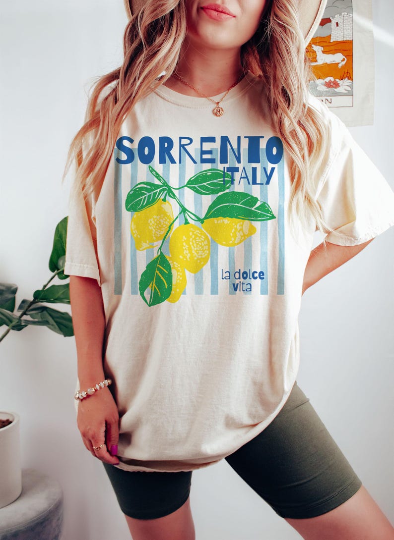Puede incluir: Camiseta color crema con las palabras "SORRENTO ITALY" en azul, con un gr&aacute;fico de rama de lim&oacute;n. La camiseta tambi&eacute;n tiene la frase "la dolce vita" en azul. El dise&ntilde;o est&aacute; sobre un fondo de rayas verticales azul claro.