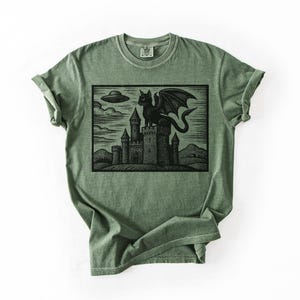 May include: A sage green t-shirt featuring a black graphic of a dragon perched atop a castle, with a UFO in the sky. The design has a vintage, woodcut style. The shirt has a classic crew neck and short sleeves.
