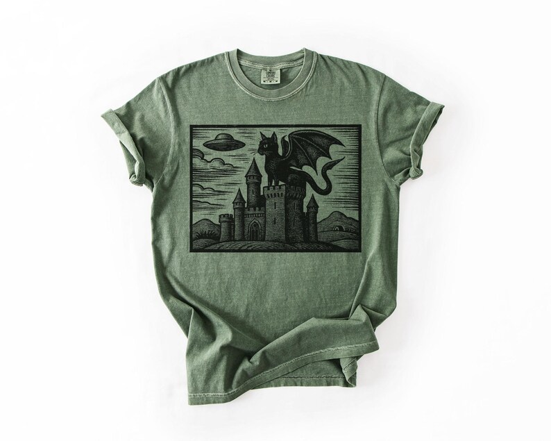 May include: A sage green t-shirt featuring a black graphic of a dragon perched atop a castle, with a UFO in the sky. The design has a vintage, woodcut style. The shirt has a classic crew neck and short sleeves.