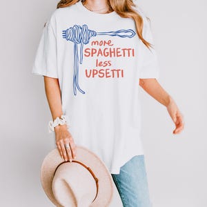 May include: White oversized t-shirt with blue and red text that reads "more spaghetti less upsetti". The text is accompanied by a blue illustration of a fork with spaghetti.