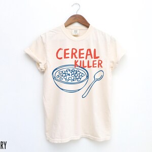 May include: A white t-shirt with a blue and red graphic of a bowl of cereal and a spoon. The text "Cereal Killer" is printed in red on the shirt.