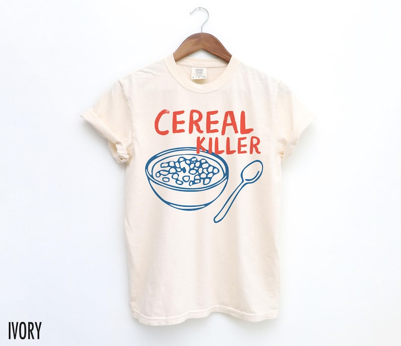 May include: A white t-shirt with a blue and red graphic of a bowl of cereal and a spoon. The text "Cereal Killer" is printed in red on the shirt.