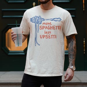 May include: A person wearing a cream coloured t-shirt with a blue and red graphic of spaghetti and the text "more spaghetti less upsetti".