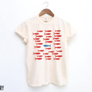 May include: A white t-shirt with a graphic of red fish with one blue fish in the middle. The text "IVORY" is in the bottom left corner of the image.