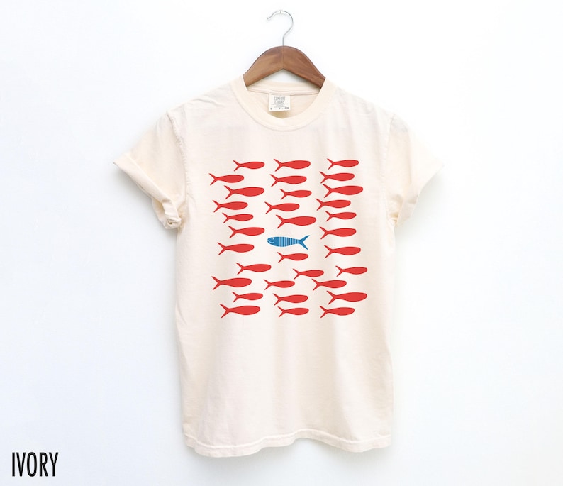 May include: A white t-shirt with a graphic of red fish with one blue fish in the middle. The text "IVORY" is in the bottom left corner of the image.