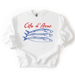 K&ouml;nnte beinhalten: Wei&szlig;es Sweatshirt mit dem Text "C&ocirc;te d'Azur" in roter Schrift &uuml;ber drei blauen Fischillustrationen. Das Sweatshirt hat lange &Auml;rmel und einen Rundhalsausschnitt.