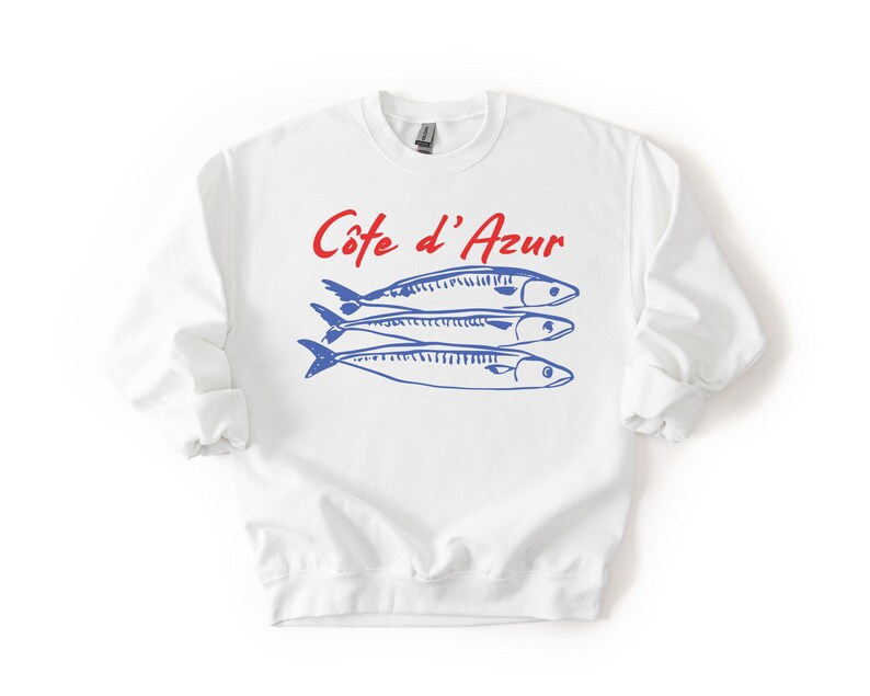 K&ouml;nnte beinhalten: Wei&szlig;es Sweatshirt mit dem Text "C&ocirc;te d'Azur" in roter Schrift &uuml;ber drei blauen Fischillustrationen. Das Sweatshirt hat lange &Auml;rmel und einen Rundhalsausschnitt.