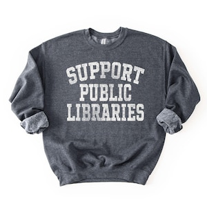 May include: Dark gray sweatshirt with the text "SUPPORT PUBLIC LIBRARIES" in white, distressed lettering. The sweatshirt has a crew neck and cuffed sleeves. The image is on a white background.