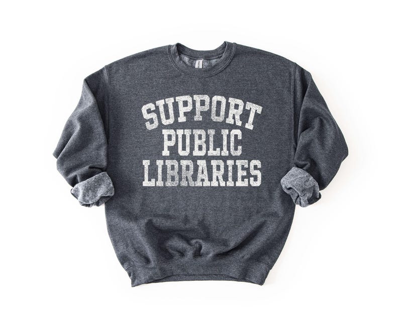May include: Dark gray sweatshirt with the text "SUPPORT PUBLIC LIBRARIES" in white, distressed lettering. The sweatshirt has a crew neck and cuffed sleeves. The image is on a white background.