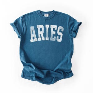 May include: A blue t-shirt with the word "ARIES" printed in white, distressed lettering. The shirt has a crew neck and short sleeves. The fabric appears to be soft and comfortable.