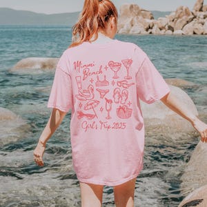May include: Pink t-shirt with a graphic design of a beach scene and the text "Miami Beach Girl's Trip 2025".