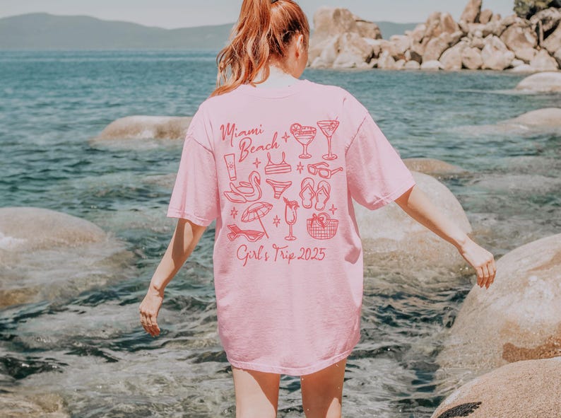 May include: Pink t-shirt with a graphic design of a beach scene and the text "Miami Beach Girl's Trip 2025".