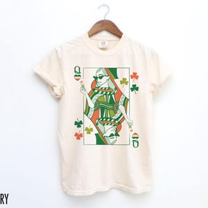 May include: Ivory t-shirt featuring a green, orange and white illustration of a queen of hearts playing card with shamrocks.