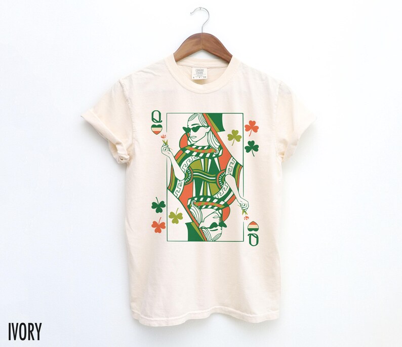 May include: Ivory t-shirt featuring a green, orange and white illustration of a queen of hearts playing card with shamrocks.