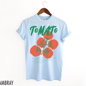 May include: Light blue t-shirt with a green "TOMATO" graphic and a drawing of red tomatoes on a vine. The text below the tomatoes reads "Their very soul. We're to the whole world. If we help will make the whole world off the page. The deliciousness is not to the fussy, unseasoned fruit."
