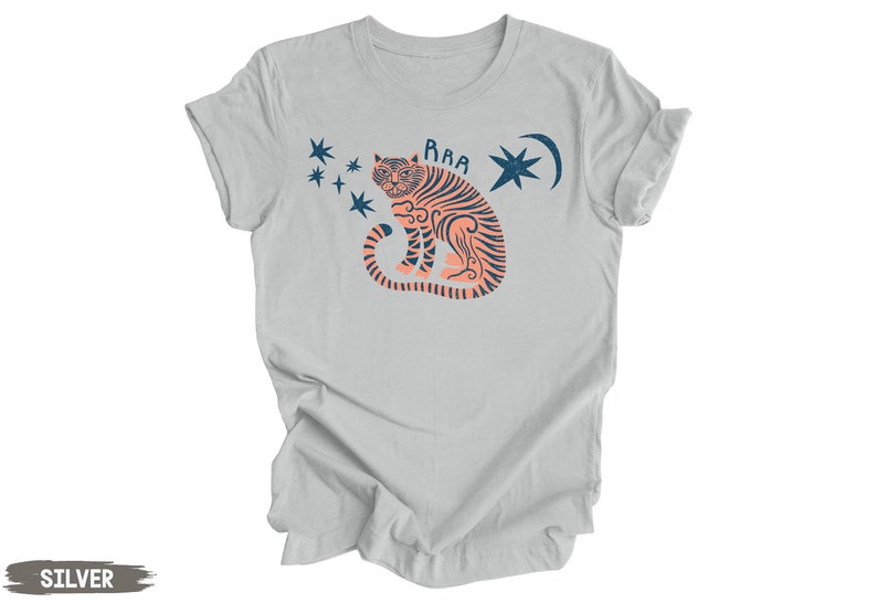 Peut inclure: Un t-shirt gris clair avec un motif de tigre rose &agrave; rayures bleues, une lune croissante et des &eacute;toiles. Le texte "RAA" est au-dessus du tigre.