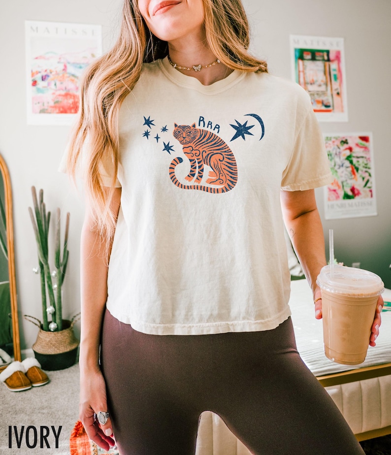 May include: Ivory-colored t-shirt featuring a seated tiger illustration in orange and navy blue, with the word "RAA" above the tiger. The design includes stars and a crescent moon. The shirt is part of a casual wear collection.