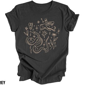 Puede incluir: Una camiseta gris oscuro jaspeada con un dise&ntilde;o gr&aacute;fico de una serpiente, estrellas y otros s&iacute;mbolos m&iacute;sticos en color marr&oacute;n claro.
