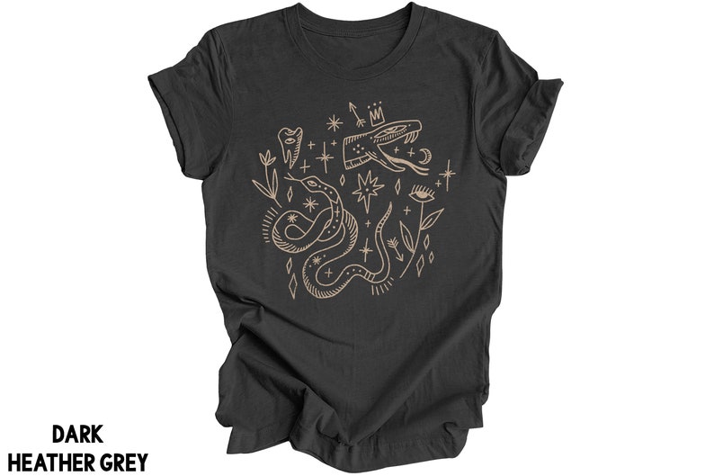 Puede incluir: Una camiseta gris oscuro jaspeada con un dise&ntilde;o gr&aacute;fico de una serpiente, estrellas y otros s&iacute;mbolos m&iacute;sticos en color marr&oacute;n claro.