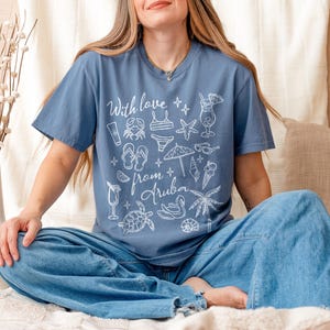 Puede incluir: Una camiseta azul con el texto "With love from Aruba" e ilustraciones blancas con tem&aacute;tica playera. La persona lleva vaqueros azules y est&aacute; sentada sobre una superficie beige.