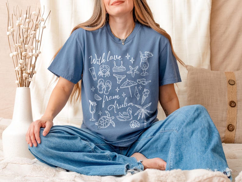 Puede incluir: Una camiseta azul con el texto "With love from Aruba" e ilustraciones blancas con tem&aacute;tica playera. La persona lleva vaqueros azules y est&aacute; sentada sobre una superficie beige.