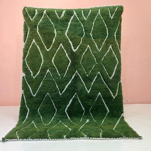 Puede incluir: Una alfombra verde con un patrón de diamantes blancos. La alfombra tiene una apariencia texturizada y afelpada y un fleco en el borde inferior. El diseño presenta formas de diamantes blancos repetidas sobre un fondo verde. Adecuado para la decoración del hogar.