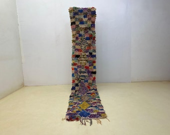 May include: A long, rectangular, hand-woven rug with a vibrant, multi-colored design. The rug features a patchwork pattern with various colors including blue, purple, yellow, and red. The rug has a textured surface and a fringe at the bottom.
