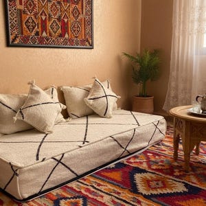 Puede incluir: Un cojín de suelo color crema con líneas geométricas negras y cojines a juego. Una pequeña mesa de madera con un juego de té y una alfombra colorida. Una obra de arte enmarcada cuelga de la pared.