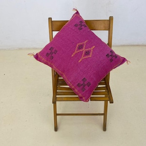 May include: A magenta square pillow with embroidered geometric patterns in black and gold, resting on a wooden chair. The pillow has tassels on the corners and a gold border. The chair is made of light brown wood.