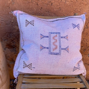 Peut inclure: Un coussin carré doté d'une base en tissu rose clair avec des motifs géométriques brodés en bleu foncé et orange. Le design comprend un motif rectangulaire central avec une ligne en zigzag et des symboles symétriques dans les coins.