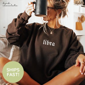 May include: A dark chocolate brown long-sleeve sweatshirt featuring the word "libra" printed in white gothic-style lettering across the center of the chest. The garment has a crew neckline and a relaxed fit, made of a solid-colored fabric.