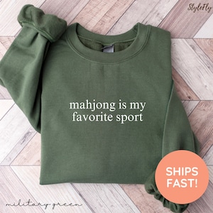 May include: Military green sweatshirt with the text "mahjong is my favorite sport" in white. The sweatshirt is a crew neck style and has long sleeves. The image also includes the text "StyleFly" and "SHIPS FAST!"