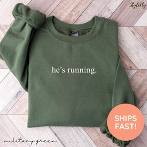 May include: Military green sweatshirt with the text "he's running" in white. The sweatshirt is a crew neck style. The words "military green" are written in cursive at the bottom of the image.