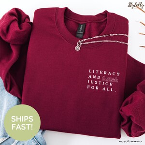 Puede incluir: Sudadera granate con el texto "Literacy and Justice for All". Un collar plateado con un colgante de sol descansa sobre la camisa. La sudadera est&aacute; doblada, con una parte de unos vaqueros visibles.