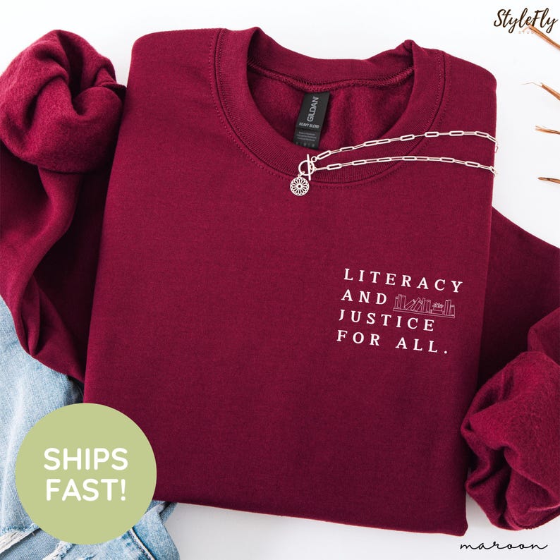 Puede incluir: Sudadera granate con el texto "Literacy and Justice for All". Un collar plateado con un colgante de sol descansa sobre la camisa. La sudadera est&aacute; doblada, con una parte de unos vaqueros visibles.