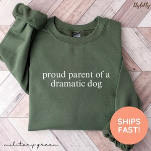 May include: Military green sweatshirt with the text "proud parent of a dramatic dog" in white. The sweatshirt has a crew neck and long sleeves. The words "military green" are written in cursive at the bottom left.