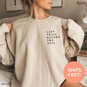May include: Sand-colored sweatshirt with the text "LAST TRAIL BEFORE THE VEIL" printed in black. The sweatshirt has a crew neck and long sleeves. The image also includes a round orange sticker that says "SHIPS FAST!"