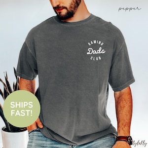 May include: A dark gray t-shirt with the text "Gaming Dads Club" in a white, curved design. The shirt is short-sleeved and has a slightly faded appearance. The word "pepper" is written in cursive in the upper right corner.