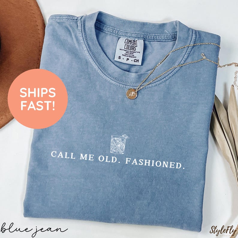 May include: A blue jean colored t-shirt with the text "CALL ME OLD. FASHIONED." and a graphic of a cocktail. A gold necklace with a pendant is draped over the shirt. The shirt has a Comfort Colors tag.