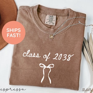 May include: Espresso-colored t-shirt with the text "class of 2038" and a bow graphic. The shirt has a crew neck and short sleeves. A silver necklace with a pendant is draped across the shirt. The shirt is folded and displayed on a white surface.