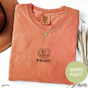 May include: A coral-colored Comfort Colors t-shirt with a pretzel graphic and the word "PROST" printed below. A gold necklace with a pendant rests on the shirt. A brown hat is visible in the upper left corner.