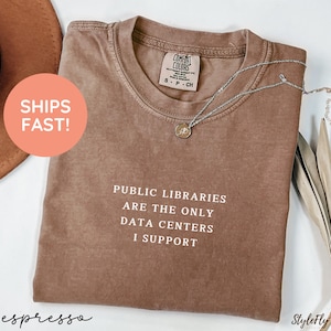May include: A brown short-sleeved t-shirt with a crew neckline and a Comfort Colors label. The shirt features white text that reads: PUBLIC LIBRARIES ARE THE ONLY DATA CENTERS I SUPPORT. A silver-toned chain necklace rests on the collar area.