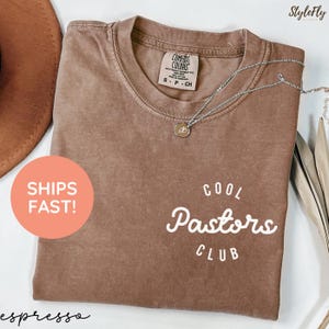 May include: Espresso-colored t-shirt with the text "Cool Pastors Club" in white script. A silver necklace with a pendant rests on the shirt. The shirt has a Comfort Colors tag.