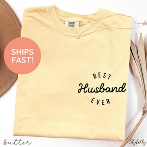 May include: A pale yellow short-sleeve t-shirt with a crew neckline. The shirt features black text on the front right side that reads BEST Husband EVER. A small Comfort Colors brand label is visible inside the collar area of the garment.