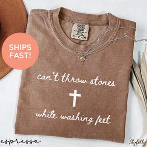 May include: A brown short-sleeved t-shirt with a crew neckline. The shirt features white text that reads "can't throw stones" above a white cross symbol, followed by the text "while washing feet" below the cross. The garment has a tag.
