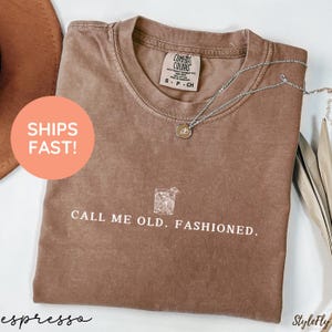 May include: Espresso-colored t-shirt with the text "CALL ME OLD. FASHIONED." and a graphic of a cocktail. The shirt has a crew neck and short sleeves. A silver necklace with a pendant is draped across the shirt.
