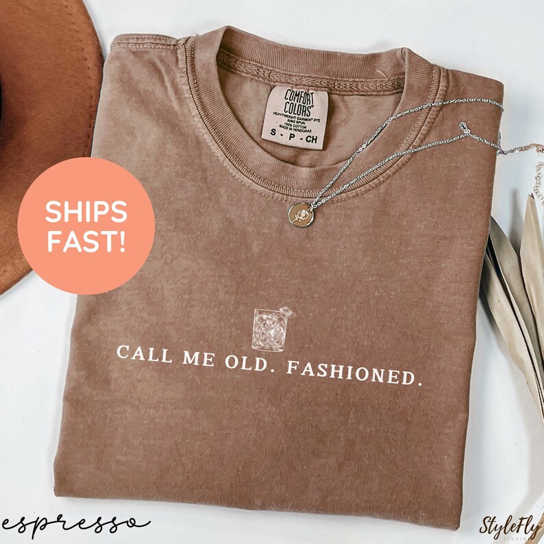 May include: Espresso-colored t-shirt with the text "CALL ME OLD. FASHIONED." and a graphic of a cocktail. The shirt has a crew neck and short sleeves. A silver necklace with a pendant is draped across the shirt.