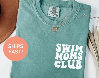 May include: A light green t-shirt with the text "Swim Moms Club" in white. The shirt has a crew neck and short sleeves. A silver necklace with a pendant is draped on the shirt. The shirt is made of a soft material.