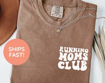 May include: Espresso-colored t-shirt with the text "Running Moms Club" in white. The shirt has a crew neck and short sleeves. A silver necklace with a pendant is draped across the shirt.