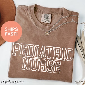 May include: Espresso-colored t-shirt with the words "PEDIATRIC NURSE" in white block letters. The shirt has a crew neck and a small tag near the collar. A silver necklace with a pendant is draped across the shirt.